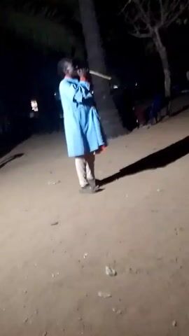 A person wearing a light blue robe and light pants stands on a sandy surface at night. The person is facing away from the camera and appears to be holding something up to their mouth. There is a palm tree to their left, casting a long shadow. In the background, several people are sitting on chairs. The scene is dimly lit.