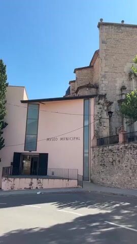 The video pans across a street in front of a building that reads "MUSEO MUNICIPAL". The building is a mix of modern and older architecture, with a light pink facade, large glass windows, and an adjacent stone structure that appears to be a church. A stone wall borders the property, topped with a fence. Several trees are visible, with lush green foliage. The sky is clear and blue, with the sun shining brightly. The street in front of the buildings is paved and shows shadows from the trees.