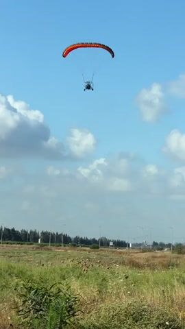 The video shows a paramotor flying in the sky. The paramotor has a red and orange wing and a pilot in a harness suspended below. The paramotor is flying over a field of dry grass and green vegetation. In the background, there is a line of trees and a blue sky with white clouds. The paramotor is moving from right to left in the frame. The video is shot from a low angle, looking up at the paramotor. There are no people visible on the ground.