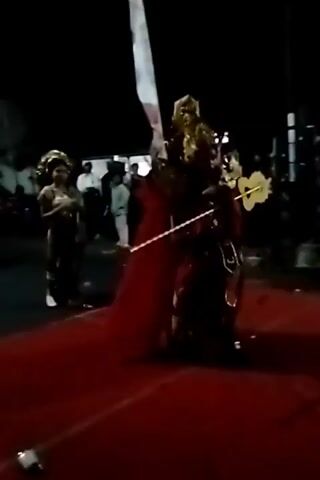 The video shows a nighttime outdoor event. In the foreground, a person in elaborate, ornate costume, possibly a performer, stands on a red carpeted area. They are wearing a golden headdress, a gold and red garment, and have long dark hair. A tall, thin white object, possibly a pole or flagstaff, is visible to the left of this performer. In the background, other people are visible, some standing and others walking. There are lights and structures in the background, suggesting a festival or performance setting. The lighting is dim, with some spotlights illuminating the performers. The camera is slightly shaky, indicating it might be handheld.