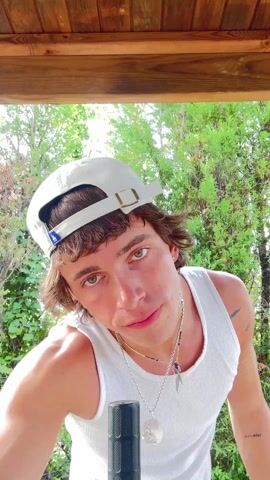 A young man in a white tank top and white baseball cap is outdoors, apparently about to sing or speak into a microphone. He stretches his arms above his head and clasps his hands together, then bends to his right, then to his left, as though warming up his muscles. The background is a wall of green foliage. The video is shot from below, with a wooden roof overhead.