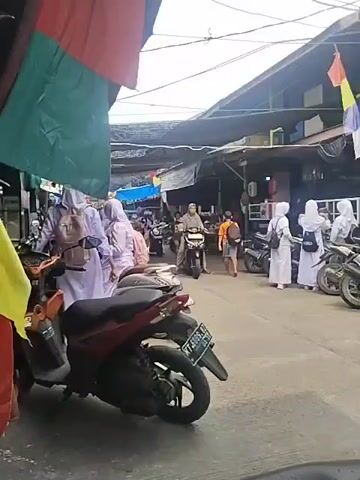 The video shows a street scene with several motorcycles and people. In the foreground, a red and black motorcycle is parked. Further down the street, a group of people, some wearing white hijabs and outfits, are seen walking and riding motorcycles. There are also market stalls or shops lining the street, covered by awnings and tarps. The overall impression is that of a busy, everyday scene in what appears to be a market or residential area.