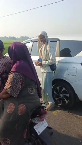 A person wearing a light-colored hijab and a beige shirt is standing near a white car with the trunk open. They are holding a small object in their hands. The car is parked on the side of a road, with a green field in the background. A food stall is set up next to the car, with signs advertising drinks like "Es Teller Kekinian" and various flavors. Another person, an older man wearing a plaid shirt, walks past the stall. The scene appears to be outdoors during the daytime, possibly in a rural area.