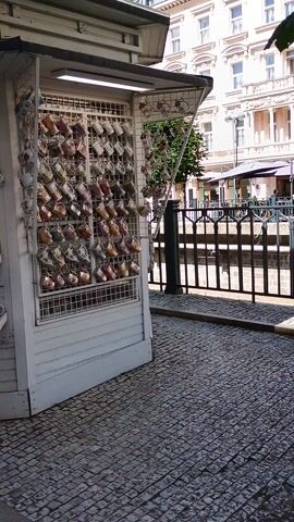 This video shows a close-up of a small souvenir shop. The shop is white and wooden, with shelves and display racks filled with various ceramic items, primarily mugs and teacups. Many of the items are adorned with intricate blue and white patterns, a style often associated with Czech porcelain. Other items on display include small decorative plates and what appear to be miniature teapots.

The shop is set up outdoors, with a cobblestone path visible to the right. There's a sense of a bright, sunny day, suggested by the lighting and the lack of visible shadows that would indicate late evening or early morning. The shop appears to be situated in a bustling area, as suggested by the cobblestones and the glimpse of buildings in the background, which are typical of European city streets. The focus is on the merchandise, with no people visible in the immediate frame, giving the impression of a quiet moment to admire the wares. The overall scene evokes a sense of traditional craftsmanship and the tourist-oriented nature of the location. The abundance of delicate ceramic items suggests a focus on collectibles and gifts.