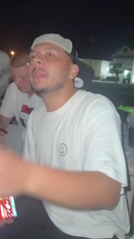 The video opens on a person wearing a white t-shirt and a trucker hat, holding a phone. They gesture with their right hand as they speak. To their left, another person in a white t-shirt looks towards the phone. A third person, also in a white shirt, is partially visible to the left of the frame. The background appears to be outdoors at night, with buildings and streetlights visible. The person in the foreground continues to talk and hold their phone, displaying what appears to be music or a video.