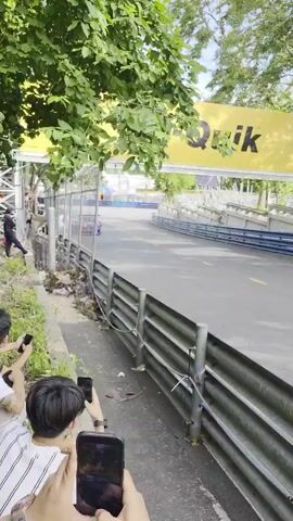 The video shows a race car speeding around a track. There are spectators on the side of the track, some of whom are filming the race with their phones. The race car is blue with orange accents and has the number 88 on its side. There is a large yellow banner with the word "Quik" on it in the background. The track is surrounded by a metal barrier, and there are trees and bushes on either side of the track. The weather appears to be overcast.