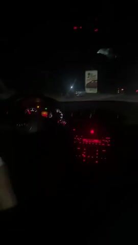 The video is shot from the perspective of someone inside a car at night. The dashboard lights, primarily red, are illuminated, showing the speedometer and radio controls. The car appears to be in motion, as suggested by the blurred lights and dark environment outside the windshield. Streetlights and distant buildings are visible. The atmosphere is dark and the focus is on the interior of the car and the road ahead.