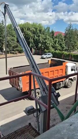 A dump truck with an orange bed is parked next to a loading platform. A crane arm extends over the truck bed, and a silver car is parked in the background. The sky is cloudy.
