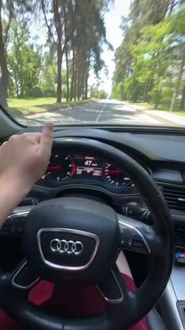 This is a first-person perspective video taken from inside a car, likely an Audi, as indicated by the steering wheel emblem. The driver's hand is visible on the steering wheel, suggesting they are operating the vehicle. The car is traveling on a paved road lined with tall, mature trees, primarily pines, creating a canopy effect. On the right side of the road, residential buildings, appearing to be apartment blocks, are visible through the trees. The road itself is a two-lane asphalt surface with a visible white line marking the center.

The car's dashboard instruments are prominently displayed, showing a speed of 49 km/h at the beginning of the clip, which then fluctuates slightly, reaching up to 47 km/h. There is also text on the dashboard display, some of which appears to be in Cyrillic script. One visible phrase translates to "God's path" or "God's way," and another reads "Speed." The overall environment suggests a sunny day with clear skies, as indicated by the bright light and shadows. The setting appears to be a suburban or semi-rural area, possibly on the outskirts of a town or city, given the presence of both natural scenery (trees) and urban development (buildings). The activity is simply driving along this road. There are no other people explicitly visible within the frame, apart from the driver implied by the first-person viewpoint. The mood is neutral, focused on the act of driving. The location context is Sarny, Ukraine.