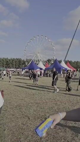 The media file depicts a bright, sunny day at an outdoor event, likely a festival. In the background, a large Ferris wheel dominates the sky, its white spokes stark against the blue. In front of the Ferris wheel, a row of colorful tents, predominantly blue, white, and red, are set up, suggesting vendor stalls or activity areas. A tall, thin black structure, possibly a flagpole or part of a larger ride, rises sharply to the right of the Ferris wheel.

The foreground and midground are populated with numerous people milling about in a grassy field. They are dressed casually, some in t-shirts and shorts, indicating warm weather. Their demeanor appears relaxed and engaged with the event. Some individuals are walking, while others seem to be congregating near the tents. The overall atmosphere suggests a lively and enjoyable gathering.

Visible text on a sign reads "MAIN STAGE" in large lettering, clearly identifying one of the main attractions of the event. Another sign, partially obscured, shows a QR code and the word "SCHEDULE". The presence of these signs and the general setup points to a structured event with organized stages and information dissemination.

The time of day is clearly daytime, with bright sunlight casting shadows. The clear blue sky with scattered white clouds suggests fair weather, contributing to the positive and energetic mood of the scene. While specific emotions are hard to gauge definitively, the general impression is one of a festive and social occasion. The combination of the Ferris wheel, tents, and numerous attendees in an open field strongly suggests a fairground or festival setting, consistent with the context of Neustadt-Glewe, Germany.