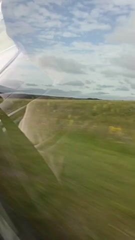 A first-person view from inside a vehicle shows a blurred landscape rushing past. The scene is dominated by green fields and a cloudy sky. The side of the vehicle, possibly a car or train window, is visible in the foreground, creating reflections and distortions of the outside view. The road or track the vehicle is traveling on is also visible, indicating motion. The overall impression is one of travel through a rural or semi-rural area.