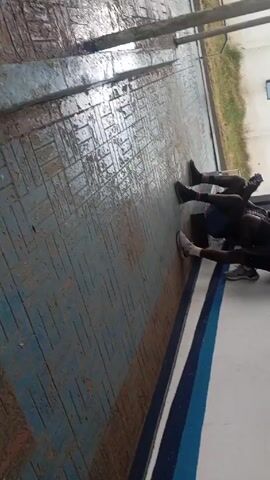 The video shows three people, who appear to be young men, on a brick surface. They are doing an exercise, lying on their backs with their knees bent and feet on the ground, performing crunches. The brick surface is blue and white with a pattern of interlocking rectangles. A white building with blue trim is visible in the background. There is also some grass and a small window visible.