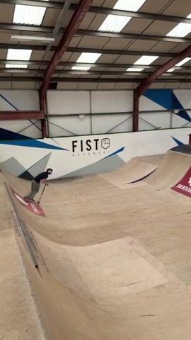 In an indoor skatepark, a person wearing a helmet and dark clothing attempts a trick on a scooter, launching into the air from a ramp. They flip upside down, lose control, and fall hard onto the ramp with the scooter landing nearby. A sign above the ramps recommends safety equipment. The park has wooden ramps with logos and words like “FIST” and “SKATEHUT.” Overhead, there are visible metal beams and bright lighting.