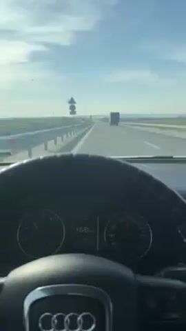 The video, shot from inside a car, shows a plane landing on a highway. The car is moving fast, as evidenced by the speedometer reading of 158-161 km/h. The plane is flying low, clearly preparing for a landing on the highway ahead. Other vehicles, including a truck and a few cars, are also visible on the road. The environment consists of a typical highway setting with roadside barriers, road signs, and an open landscape with distant hills. The weather is clear with a blue sky.