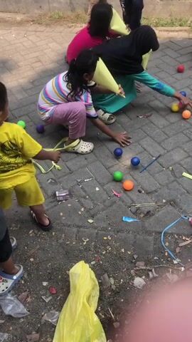 This is a top-down, slightly angled, eye-level shot of a group of children playing a game outdoors on a paved surface. The children, mostly girls, are wearing casual clothing and some have yellow cone-shaped hats or visors on their heads. They are crouching or bending down, reaching for and interacting with small, colorful balls scattered on the ground. There are blue, green, orange, and pink balls visible. Some children are using their hands to pick up or manipulate the balls, while others appear to be aiming them towards a cardboard box with a hole in it, positioned on the right side of the frame. Yellow ribbons or streamers are also spread across the ground, along with some debris like wrappers and sticks. In the background, other children are standing or walking, and part of a wall or fence is visible. The overall scene depicts a lively and active game being played by children in an outdoor setting.