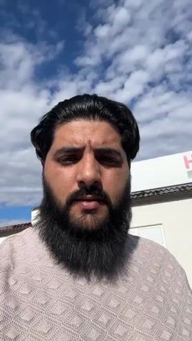A bearded man with dark hair, wearing a light-colored textured sweater, films himself outdoors in a selfie perspective. The sky is blue with white clouds. In the background, a white building with a red "HERMANN" sign and window displays featuring people engaged in outdoor activities is visible. A young boy in a blue t-shirt with a "GAMES" graphic appears next to the man, smiling and giving a thumbs-up while holding the man's hand. A woman in a light purple top walks away in the background. The scene is an outdoor paved area with some trees.