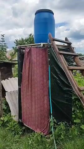 A rudimentary outdoor shower setup is shown, featuring a large blue barrel on top of a makeshift structure. The structure appears to be made of dark tarpaulin or plastic sheeting, draped over a frame. A patterned fabric curtain hangs on the front, partially obscuring the interior. A wooden ladder is leaning against the right side of the structure, providing access to the barrel. A green hose is visible hanging down the side. The scene is set outdoors with lush green grass and foliage surrounding the shower. In the background, there are hints of rural buildings and a cloudy sky. The video appears to be a static shot, with no discernible movement or action.