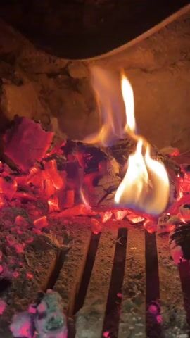 The video shows a close-up view of burning embers and a piece of wood in a fireplace. The embers are glowing red and orange, and flames are flickering around the wood. The wood appears to be partially charred and is actively burning. The fireplace grate is visible below the embers. The background is dark and suggests the interior of a fireplace or hearth. The lighting is dim, with the primary illumination coming from the fire itself. There are no people or other objects visible in the frame.