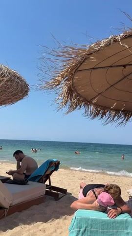 This video shows a beach scene on a clear, sunny day with a bright blue sky. Two large thatched umbrellas provide shade on a sandy beach. In the foreground, a person lies on a beach chair under one umbrella, facing away from the camera with a pink cap on their head and a dark swimsuit. Another person sits in a chair to the left, looking down at a phone, wearing dark shorts. In the background, several people are swimming in the calm, blue ocean, with a distant shoreline visible under a hazy sky. The water appears clear with gentle waves lapping the shore.