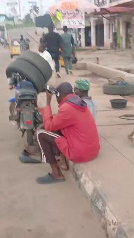 The video captures a daytime street scene in Shaba, Nigeria, focusing on a group of men and a motorcycle at the roadside.

In the foreground, two men are seated on a low concrete barrier or curb. The man on the left, wearing a red hooded jacket and dark track pants with white stripes, is engrossed in his smartphone, holding it up with both hands. He is wearing a blue slipper on one foot, while the other is bare. Beside him, to the right, sits another man in a grey shirt and a distinctive green and white patterned cap, also looking towards the phone screen.

Adjacent to these men, on the left, is a blue motorcycle, noticeably laden with several dark tires stacked high on its rear seat and luggage rack, indicating it might be used for transporting tires. The motorcycle's rear wheel, mudguard, and taillight assembly are visible.

The background shows a paved road extending into the distance. Several other individuals are walking along the street; two men are seen walking away from the camera, and another in a light-colored top is further left. A yellow auto-rickshaw (commonly known as a 'keke napep' in Nigeria) is partially visible further down the road on the left.

On the right side of the frame, alongside the road, additional tires are scattered or stacked on the ground next to a black basin, suggesting an informal tire repair or sales point. Buildings with awnings line the street in the background, contributing to an urban or semi-urban setting. The natural light indicates it is daytime. No specific text is clearly legible on any signs or objects within the frame. The overall atmosphere appears casual, depicting daily life at a roadside location.