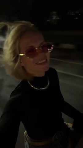 A blonde woman with short hair and pink sunglasses is riding on the back of a vehicle at night. She is wearing a black long-sleeved shirt with a sheer neckline and a silver necklace. Her head is tilted back and she is smiling. In the background, there are blurred lights from buildings and streetlights, indicating an urban environment. The motion of the vehicle is causing the image to be blurry, suggesting the footage was captured while in motion.
