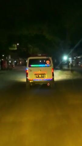 A white van is shown from behind driving down a street at night. The van has multi-colored LED lights along the rear bumper and taillights. The van also has a sticker on the back window. The street is paved and appears to be in a town or city, with buildings and trees visible on either side of the road. The lighting is dim, with streetlights and the van's lights providing illumination. There is some motion blur in the video, indicating the van is in motion.