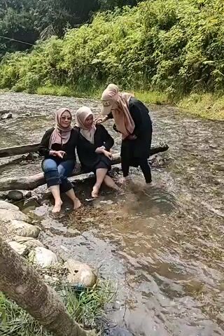 Three women are sitting and standing by a river. Two women are sitting on a log with their feet in the water. A third woman is standing in the water behind them, holding a phone. The river is shallow and clear with rocks visible on the riverbed. Lush green foliage covers the bank behind the women.