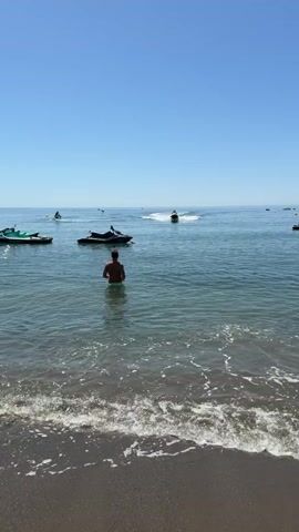 A person stands in shallow water at a beach, facing away from the camera. Several jet skis are visible in the water, some stationary and some in motion. One jet ski is in the foreground, to the left of the person. Another jet ski is further out in the water and appears to be moving. The sky is clear and blue. The water is calm with small waves breaking on the shore. The beach appears to be sandy.