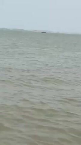 The camera is positioned at water level, looking out over a body of water with small waves. In the distance, land is visible. A small boat comes into view from the right, bobbing in the water. The boat appears to be a fishing boat with equipment on board. The sky is overcast.