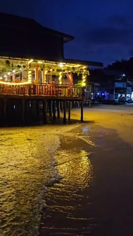 A nighttime view of a restaurant built on stilts over the water. The restaurant is lit with strings of warm yellow lights. Waves gently lap at the sandy shore in the foreground. The sky is a deep blue, indicating it is either dusk or dawn.