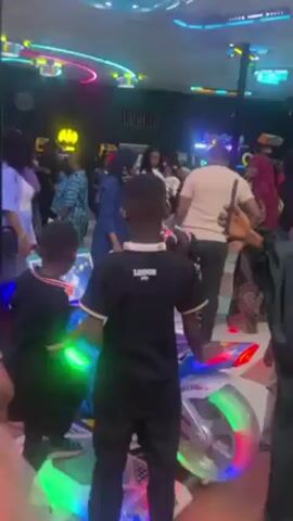 The video shows the interior of a bustling entertainment center or arcade. There are many people, including adults and children, milling around. Some are interacting with arcade games, including a motorcycle racing simulator that is flashing with neon lights. The lighting is bright, with various neon signs and overhead fixtures contributing to a vibrant, energetic atmosphere. The camera focuses on several individuals in the crowd, including a woman with curly hair, wearing a white top and a light blue shirt, who looks towards the camera. Children stand in the foreground, giving the impression they are observing the various activities.
