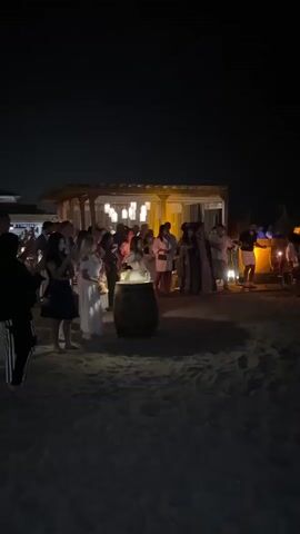 The video shows a nighttime gathering on a sandy beach. In the foreground, a dark sandy area with some footprints is visible. In the middle ground, a group of people are standing around a barrel with something lit on top, possibly a candle or a small fire. They are positioned near a low-rise structure with a thatched roof and lights illuminating it from within. Beyond this structure, more people are spread out, some seated at tables. The overall atmosphere appears to be a social event or party. The lighting is dim, with artificial lights creating a warm glow in the area of the gathering. The background is dark, suggesting it is nighttime.