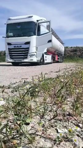 The video shows a white DAF semi-truck with its driver's side door open. The truck is parked on a dirt road. The trailer appears to be a large tank. The environment is outdoors with a blue sky and some clouds. There are some plants and rocks on the ground in the foreground.
