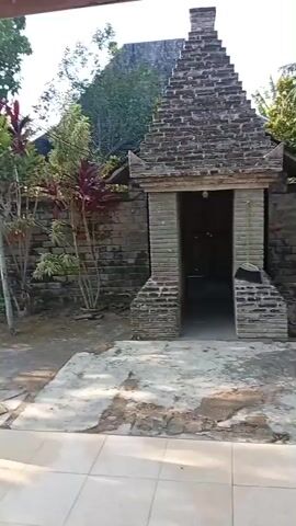 A video pans across an outdoor scene. In the foreground, a tiled patio is visible with the underside of a wooden roof overhead. The camera moves to the right, revealing a brick building with a distinctive pyramidal roof. Adjacent to this structure, a stone wall separates the yard from a densely vegetated area. Various plants, including some with large red leaves, are present. Further to the right, several small, colorful structures resembling graves are arranged in a row. A dry palm tree stands in the middle ground, and beyond it, more greenery is visible, with a hint of a larger stone structure in the background. The lighting suggests it is daytime, possibly with some sunlight filtering through. The overall impression is that of a courtyard or garden area, potentially part of a historical site or cemetery, with elements of traditional architecture and natural landscaping.