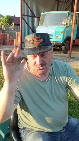 The video shows a close-up of a middle-aged man wearing a green t-shirt and a black Harley-Davidson baseball cap. He has a mustache and appears to be speaking, with his hand raised in front of him. In the background, a light blue truck is visible, along with a white van. The scene appears to be outdoors, possibly in a yard or garage, with a metal wall or fence and some grass. The lighting suggests it might be late afternoon or early evening. The camera angle is slightly angled upwards towards the man.