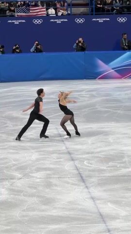 Two figure skaters, a man and a woman, are performing on an ice rink. They are both dressed in black athletic attire. The woman wears a black leotard with sheer tights, and the man wears a black long-sleeved top and black trousers. They are holding hands and skating in synchronized motion, moving across the ice. The ice surface shows several skate marks.

In the background, a large blue barrier with white and pink abstract designs is visible. Along the top of this barrier, the Olympic rings logo is displayed multiple times, alongside the text "MILANO CORTINA 2026." Above the barrier, spectators are seated in stands, and several camera operators are positioned, including one operating a large camera on a crane arm. The lighting appears bright, suggesting an indoor arena.