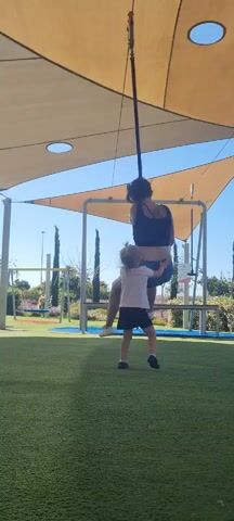 A woman in a black tank top and denim shorts sits on a horizontal bar suspended from a pole, swinging under a tan canopy. A small child in a white shirt and dark shorts stands in front of the woman, reaching up to her. The child appears to be looking up at the woman. They are on a green artificial turf surface at a playground. The sky is clear and blue. There are other playground structures and trees in the background. The woman smiles slightly.