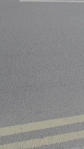 A low-angle, static shot of an asphalt surface with white road markings is visible. The markings appear to be part of a parking bay, with angled lines forming a rectangular shape. The asphalt is textured and gray. In the distance, a slight blur suggests movement, and a curb is faintly visible. The scene appears to be outdoors, possibly in a parking lot or on a street.