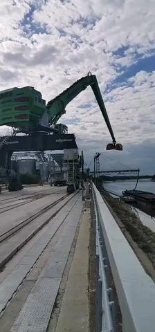 The video shows a large green industrial crane with a yellow bucket attached to its arm. The crane is positioned over a waterfront area with a river or canal. The bucket is lowered towards a barge in the water, suggesting a loading or unloading operation. In the background, there are more barges, a tugboat, and a bridge. The sky is partly cloudy. The scene appears to be a port or industrial facility.