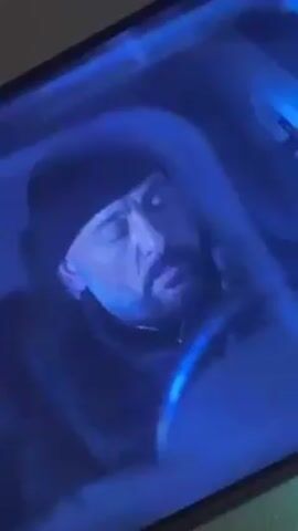 A video shows a man sitting inside a car. The man is wearing a black beanie and a black jacket. He has a beard and mustache, and he is holding a cell phone up to his ear. The interior of the car is illuminated by a blue light.