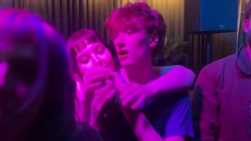 The video shows a group of young people in an indoor setting, likely a club or music venue, bathed in purple and pink neon lighting. In the center, a woman with dark hair and bangs, possibly with tattoos on her arm, holds hands with a man with curly reddish-brown hair who is wearing a blue t-shirt and has an earbud in his ear. They look engaged and possibly intimate. To the left, a woman with blue/green highlights in her hair and a nose ring frequently looks at the camera, sometimes playfully sticking out her tongue. A bearded man in a dark shirt is partially visible on the right. In the background, dark curtains, part of a drum kit, a plant, and a speaker can be seen, indicating a performance space. The atmosphere appears lively and playful, suggesting a social gathering or concert at night in Dundee, United Kingdom. No text is visible.