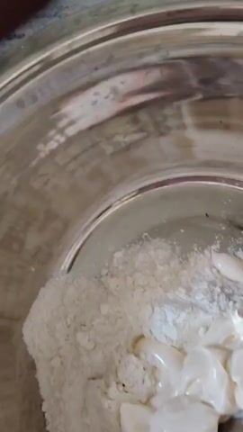 The video shows a close-up of a stainless steel mixing bowl with a white powdery substance and a dollop of a creamy white substance. A spoon is partially submerged in the creamy substance, appearing to be mixing it. The background is blurred, but hints of a kitchen environment are visible. In the next shot, a hand is seen pouring a portion of white shredded material from a clear plastic bag into a bowl, and a white bag with some text is also visible. The video then transitions to a shot of a person holding a bowl with three raw eggs in it. This bowl is then tilted over the mixing bowl, and the eggs are poured into the mixture of powdery and creamy substances. The action of pouring the eggs into the bowl is captured as they mix with the existing ingredients.