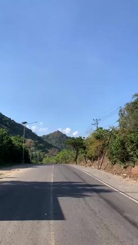 A paved road curves through a hilly, green landscape under a clear blue sky. The road is asphalt with a broken yellow line down the center and a white line on the right edge. On the left side of the road, a tall lamp post with a solar panel is visible. On the right side, trees and foliage line the road, with telephone poles and wires also present. The hills in the background are covered in trees and vegetation, with one prominent peak in the distance. The scene appears to be taken from a moving vehicle.