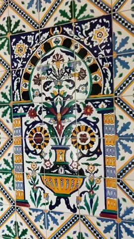 This video shows a close-up view of a decorative tiled wall with a central floral design. The tiles are arranged in a repeating pattern of diamonds and squares, featuring geometric motifs and stylized floral elements in colors such as blue, green, yellow, and white. The central design is more elaborate, depicting a vase filled with colorful flowers and foliage framed by an arched motif at the top. The lighting appears to be natural, highlighting the textures and colors of the tiles. There are no people or obvious actions occurring in the video, just a static view of the tiled wall.