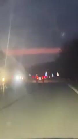 The video shows a view from inside a car driving on a road at dusk. The sky is a mix of pink and dark blue. There are other cars on the road ahead, with their headlights and tail lights visible. Trees line the sides of the road.
