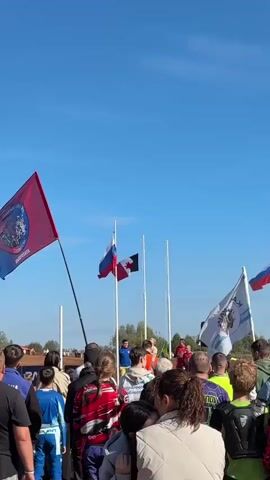 The video shows a crowd of people gathered outdoors under a clear blue sky. Several flags are visible. On the left, a large red flag with a circular logo is held on a pole. In the center, two smaller flags are displayed on poles: one is the Russian flag, and the other is a black and white flag. To the right, a white flag with a logo and text is flying, and further to the right, another flag with red and blue colors is visible. The people in the foreground are facing away from the camera, and some appear to be looking towards the flags or a central area where some individuals are standing. The environment appears to be an open field with some distant trees. The overall scene suggests an outdoor event or gathering.