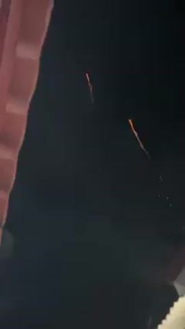 The video shows fireworks exploding in the night sky. The camera angle is looking up from what appears to be a balcony, with a pink wall visible on the left and a small portion of a white object on the bottom right. There are multiple firework explosions, some displaying sparks. The background is a dark, clear night sky.