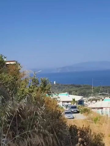 A video shows a view from a hillside overlooking a bay. In the foreground, dry grasses and green shrubs are visible. A few buildings with light-colored walls and blue-trimmed roofs are clustered on the hillside. A paved road winds among the buildings, with a few cars parked on it. In the middle ground, the blue water of the bay stretches out, with a distant island or coastline visible on the horizon. A white lighthouse is also visible on a point of land jutting into the water. The sky is clear and blue.