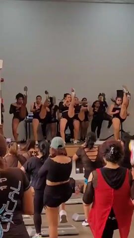 A group of women is performing a dance routine on a stage in a studio. They are all wearing black athletic wear, and some of them have their hair pulled back. The women are moving in unison, and their movements are energetic and synchronized. The studio is brightly lit, and the walls are painted a light gray. There are several rows of people in the foreground, also dressed in black, watching the performance. They are all facing the stage, and some of them are mirroring the dancers' movements.