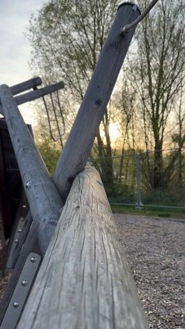 The camera focuses on a wooden structure in a playground, likely part of a climbing frame. The logs are weathered and grey, and the sun shines through the trees in the background. There's a wire fence visible in the distance. The ground is covered with wood chips. A ladder and hanging rings are attached to the structure. A child is briefly heard in the background.